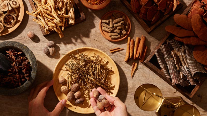 an arial view of hands holding and counting out Chinese herbs in a large group of herbs in wooden bowls, plates, and boxes on a wooden counter