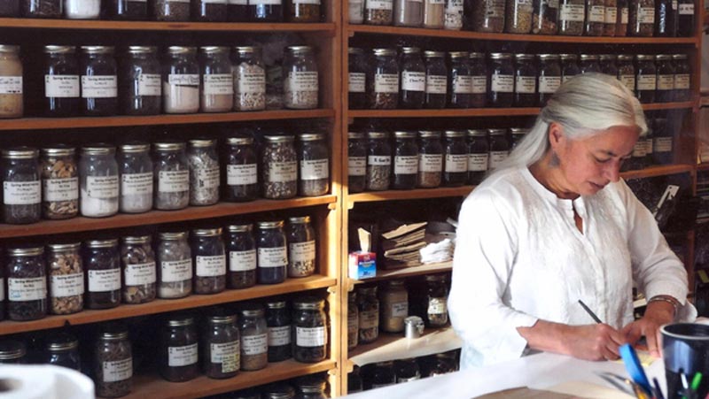 a closeup of a woman with gray hair wearing a white doctor's coat stands at wooden counter in a room of shelves with Chinese medicinal herbs writing
