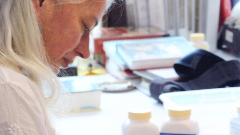 a extreme closeup of woman with gray hair wearing a white doctor's coat stands at wooden counter in a room of shelves with Chinese medicinal herbs writing